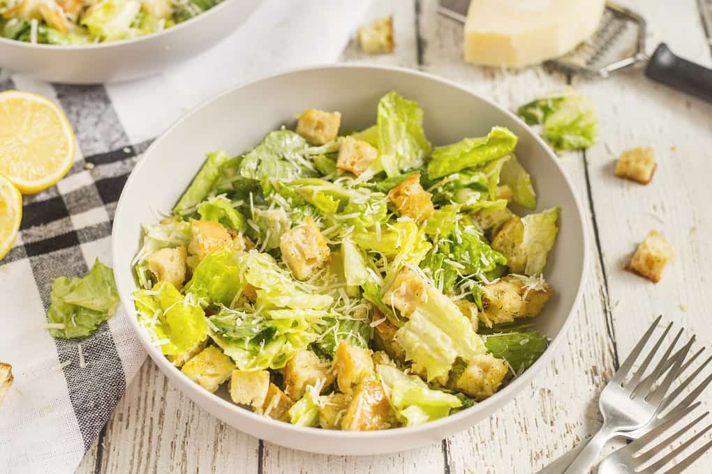 Individual white serving bowl with Caesar salad on a white board with forks