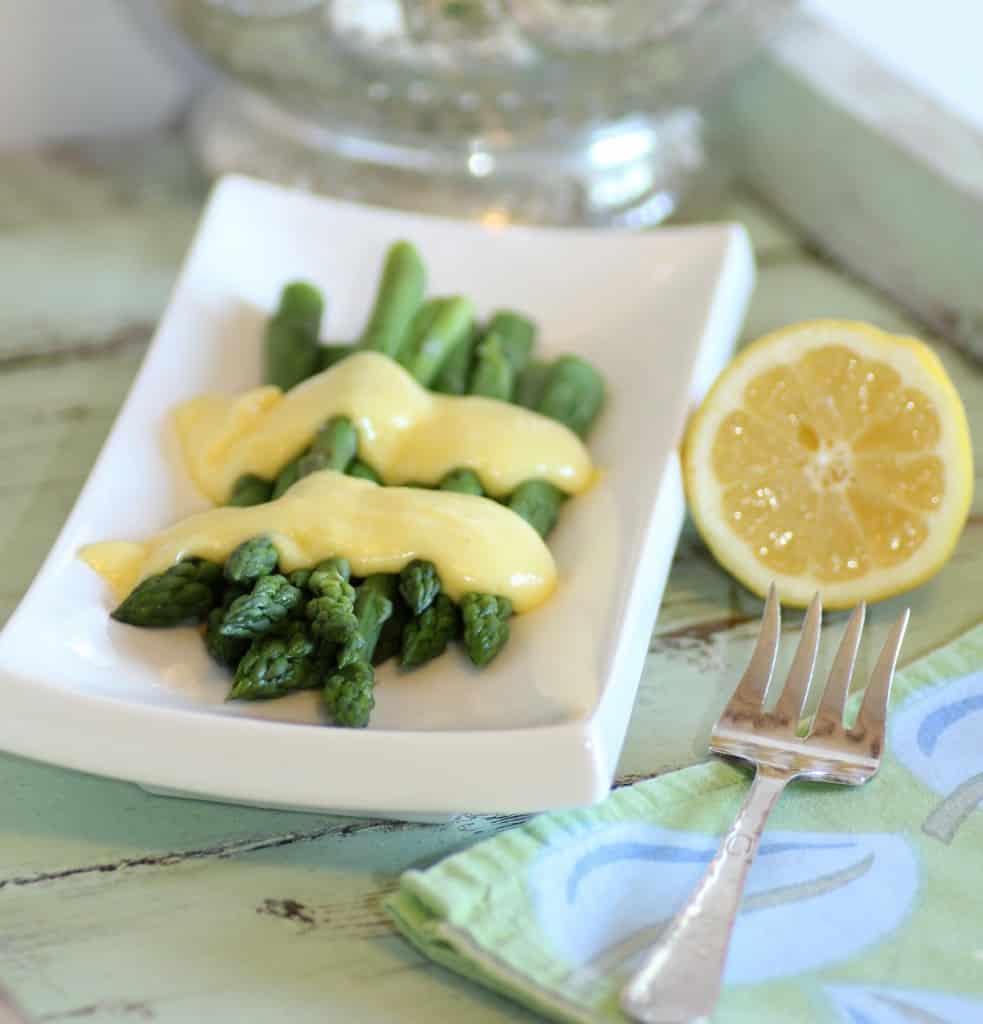 Julia Child's Asparagus with Hollandaise Sauce on a rectangle white plate with a serving fork and a 1/2 a lemon