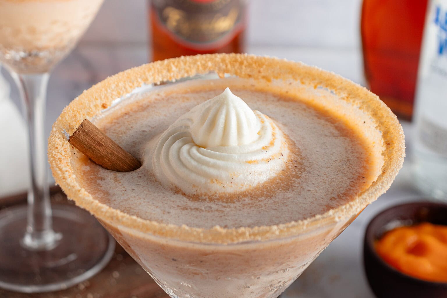 A close-up of a creamy cocktail in a martini glass with a crumb sugar rim. The drink is topped with a swirl of whipped cream and a cinnamon stick. Blurry background elements include a bottle and another cocktail glass.