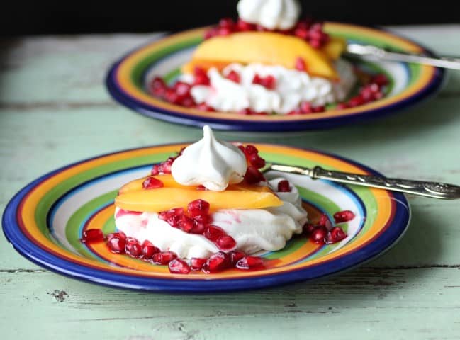Pavlova on a multi-colored plate with a slice of mango and pomegranate seeds and silver fork 