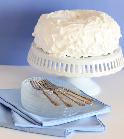 Blue Hawaiian Bundt Cake on a white cake stand with glass serving plates stacked beside it and forks on top of the plates