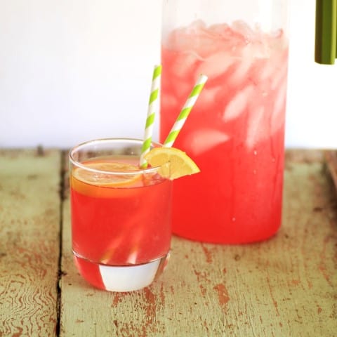 Rhubarb Lemonade in a glass with ice and a straw with a full pitcher behind