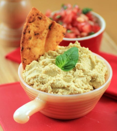 Classic Mediterranean Hummus in a white bowl with a pita chip and a basil leaf for garnish