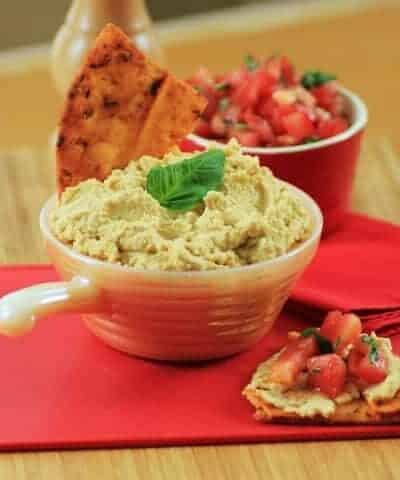 Mediterranean Hummus in a small white serving bowl with some on a cracker with tomotoes