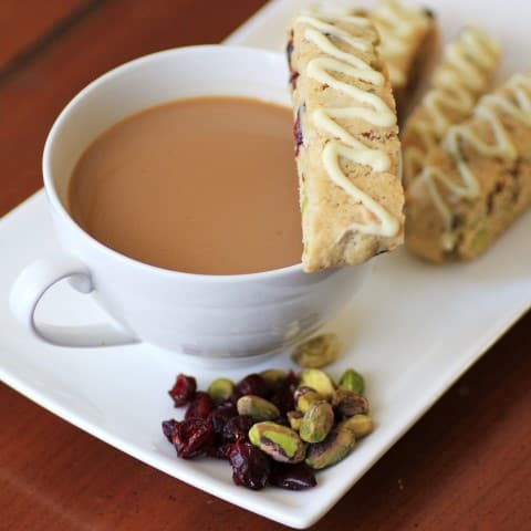 Cranberry Pistachio Biscotti sitting on the rim of a cup f coffee with dried cranberries and pistachios on a plate