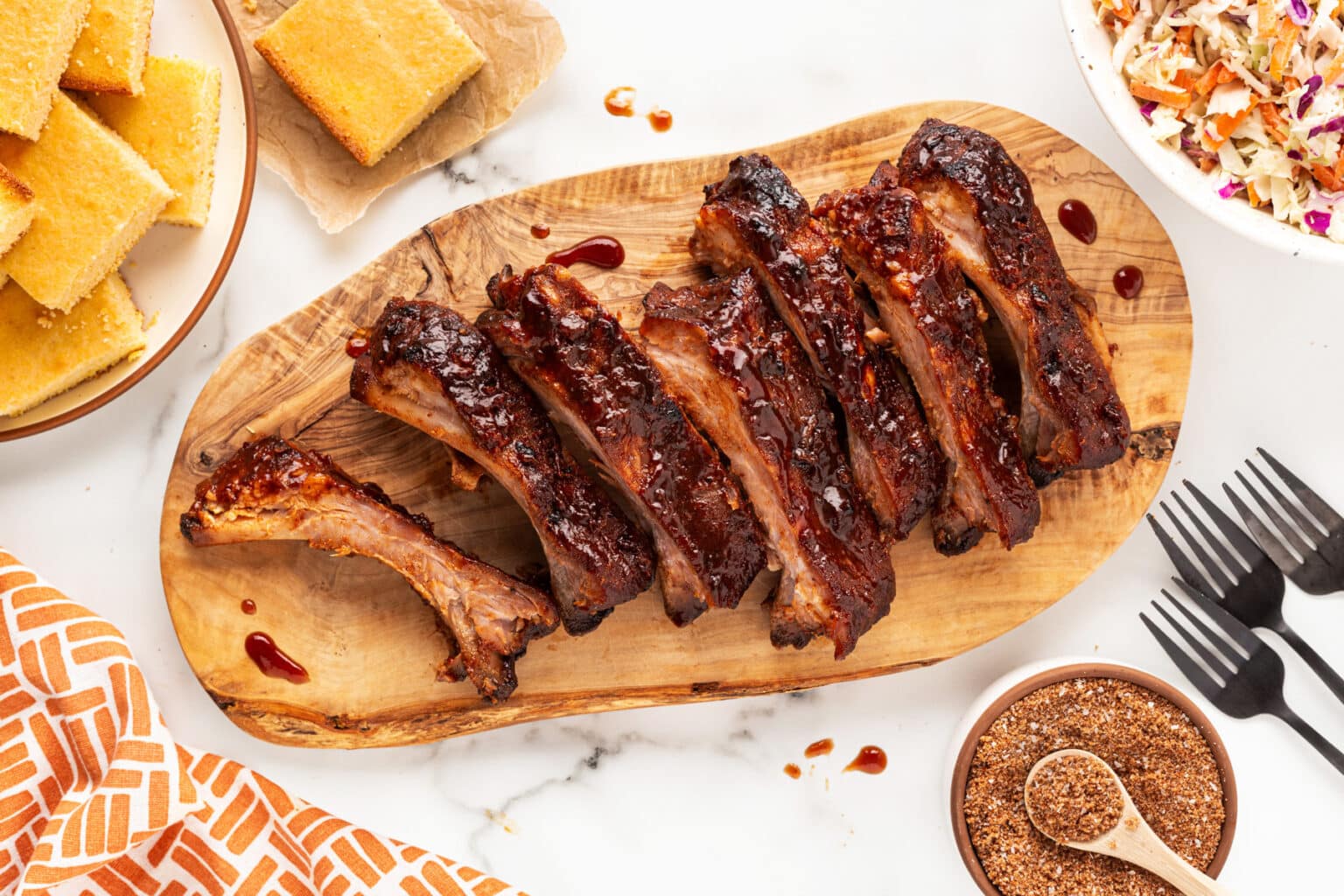 A wooden board holds a rack of glazed, cooked ribs with barbecue sauce. Around the board, there are slices of cornbread, a bowl of coleslaw, a spice bowl, three black forks, and an orange patterned napkin on a white surface.