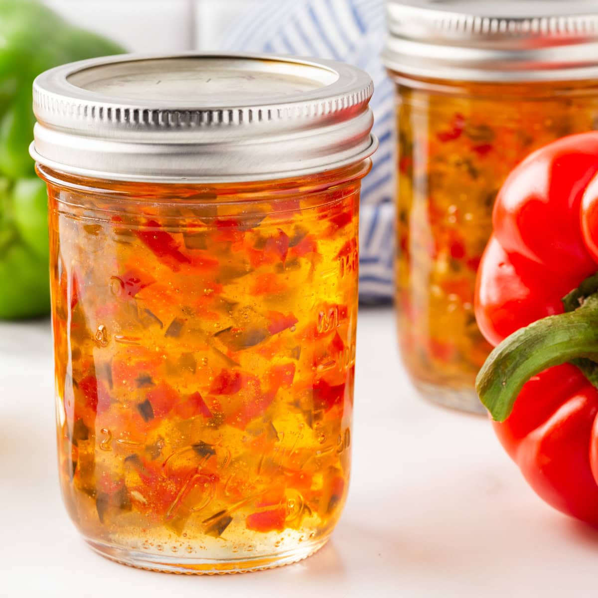 Square photo of red and green pepper jelly in a jar with a red pepper beside it. 