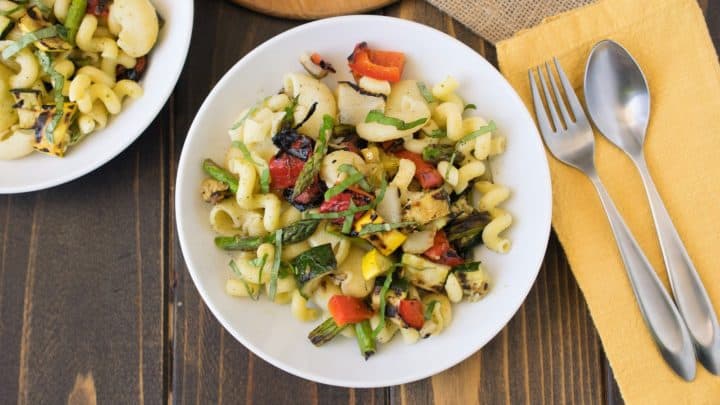 Grilled spring vegetable pasta salad in a white bowl with a fork and spoon beside the bowl