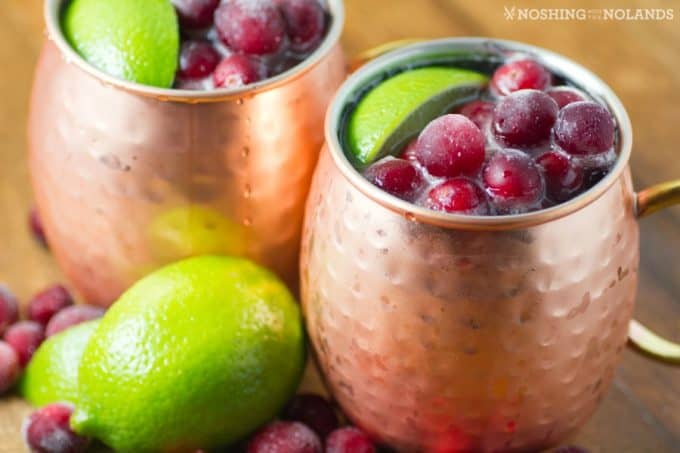 Two copper mugs filled with a dark red drink, topped with fresh cranberries and lime wedges. A few whole limes and cranberries are scattered around the mugs on a wooden surface.