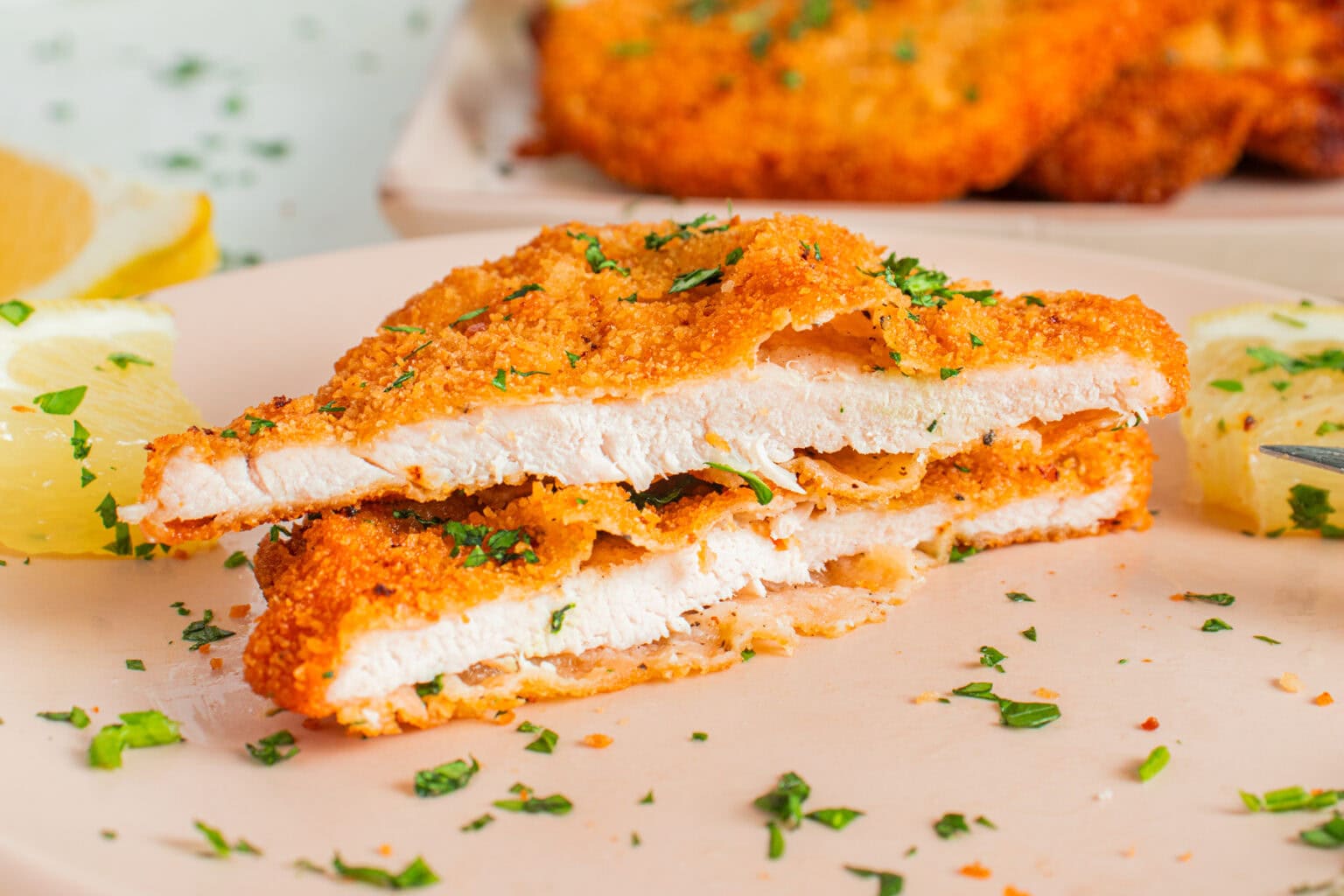 Close-up of crispy fried chicken cutlets garnished with parsley, served with lemon wedges on a pink plate.
