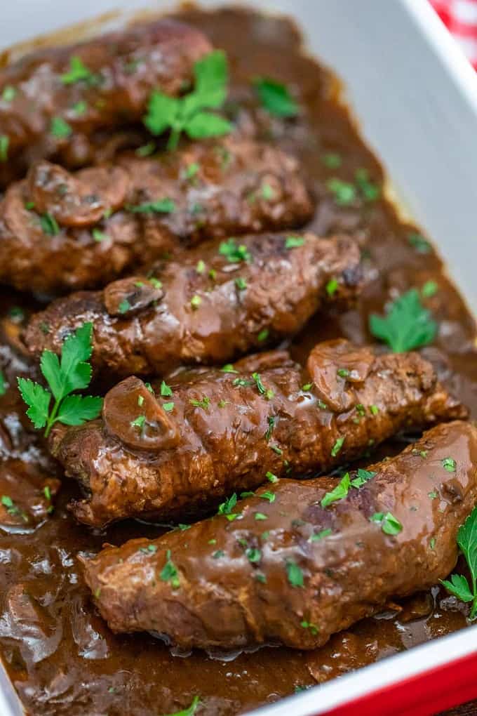 Beef rouladen in a red and white casserole dish with mushroom gravy and cilantro