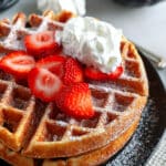 Stacked waffles on a plate with icing sugar, strawberries and whipped cream.