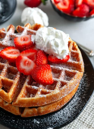 Stacked waffles on a plate with icing sugar, strawberries and whipped cream.