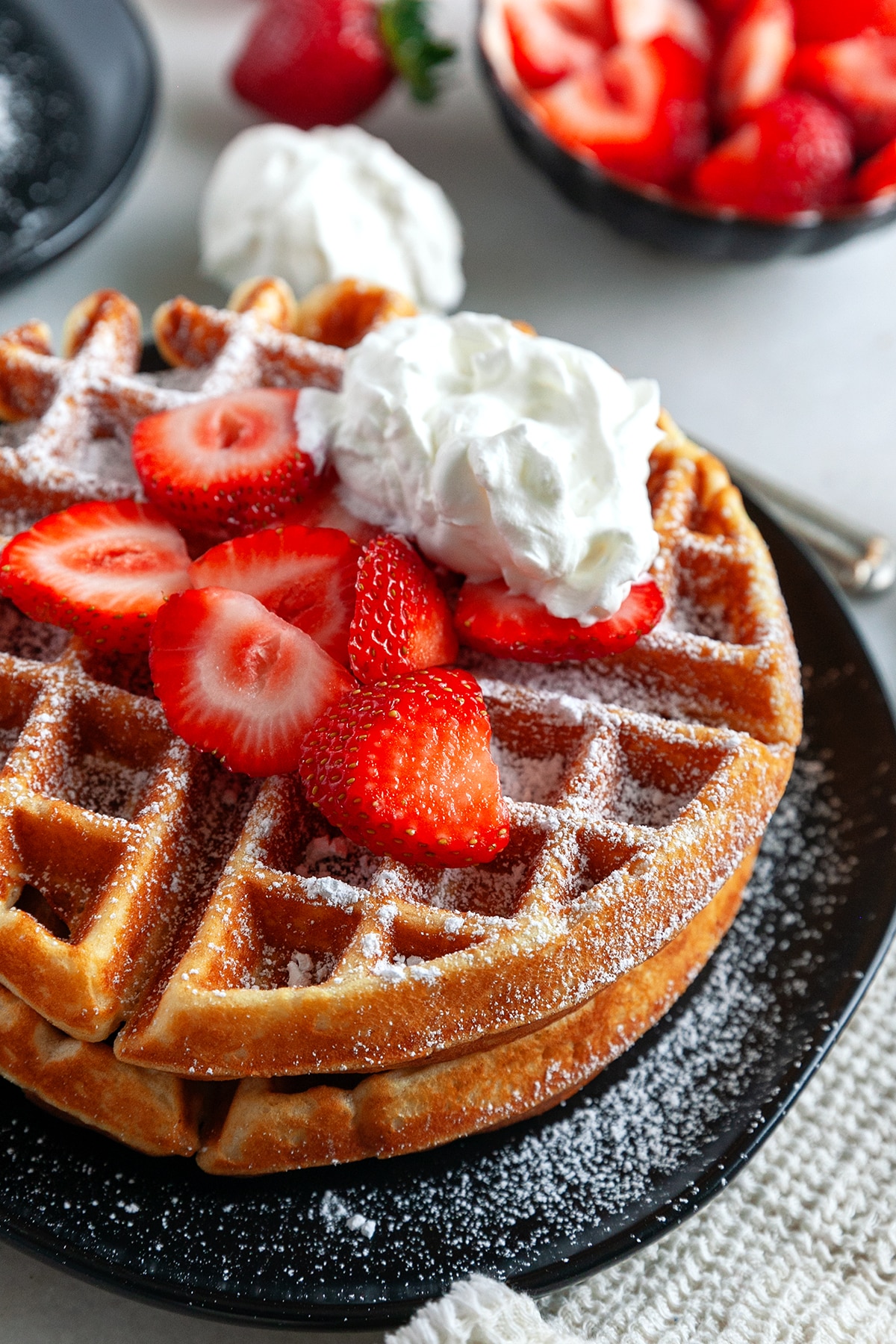 Showing a close up of the Belgian Waffle Recipe with two waffles stacked, icing sugar dusted, strawberries and whipped cream. 