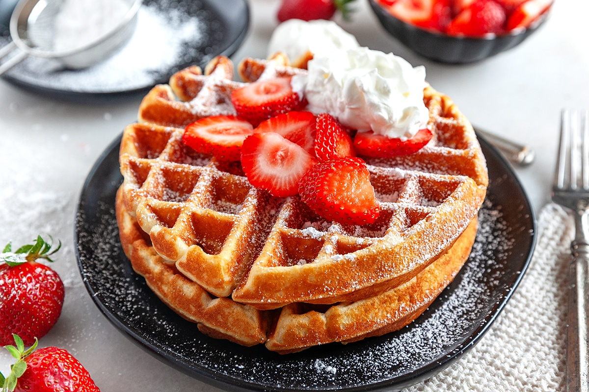 Horizontal photo of two stacked Belgian Waffles with strawberry, icing sugar and whipped cream. 
