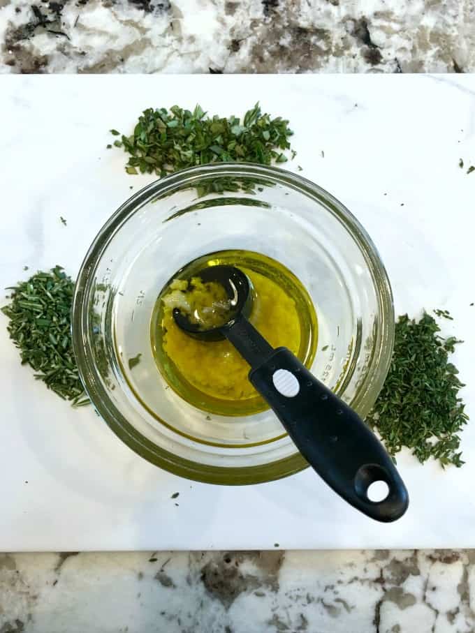 Olive oil in a small glass bowl with garlic and fresh herbs surrounding. 
