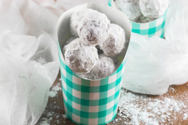 A checkered teal and white cup holds several powdered sugar-covered doughnut holes, with more in the background on a wooden surface. White cloth is draped around the scene, and a dusting of sugar is sprinkled on the table.