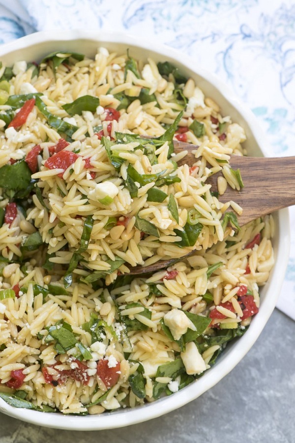 Orzo salad with roasted red peppers, spinach and feta in a white bowl with a wooden serving spoon