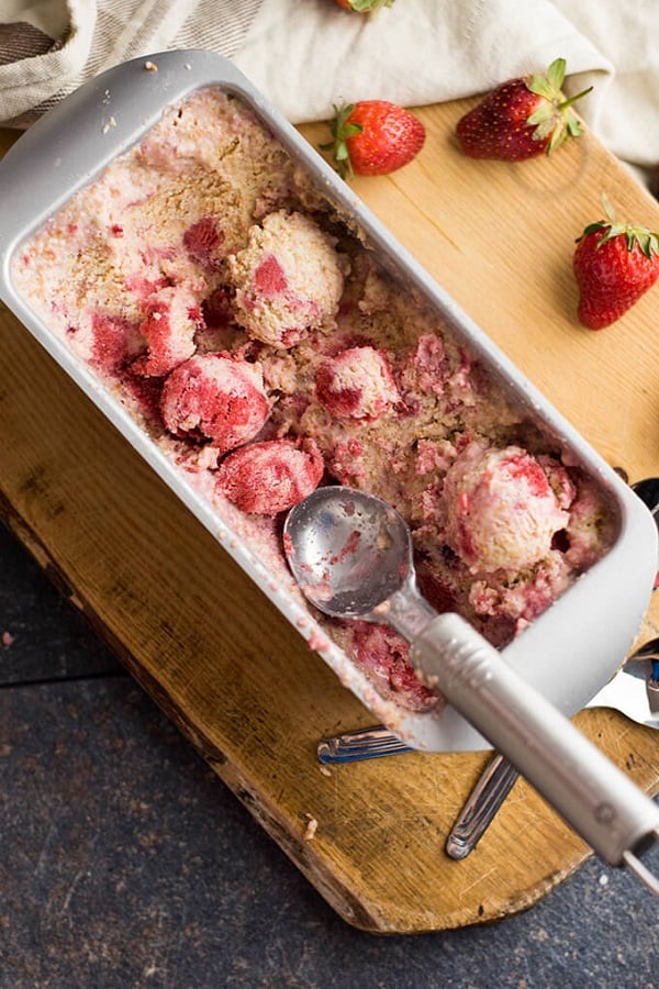 A pan of strawberry cheesecake ice cream on a wooden board with an ice cream scoop and some strawberries