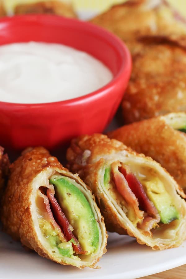 Easy Avocado Bacon Egg Rolls Recipe sliced in half on a white plate with dip in a red bowl