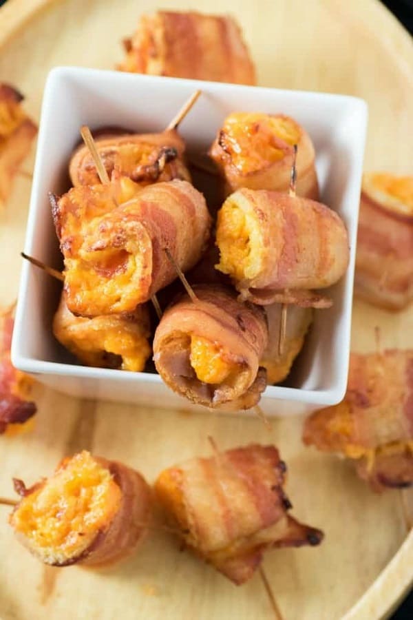 Bacon Cheddar Roll Ups in a small white bowl overflowing onto a wood tray from 18 Easy Hot Party Appetizer Recipes