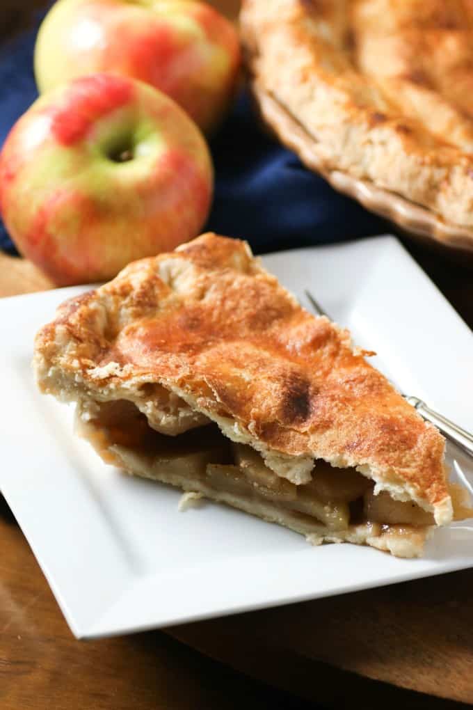 Slice of apple pie on a white plate with apples in the background