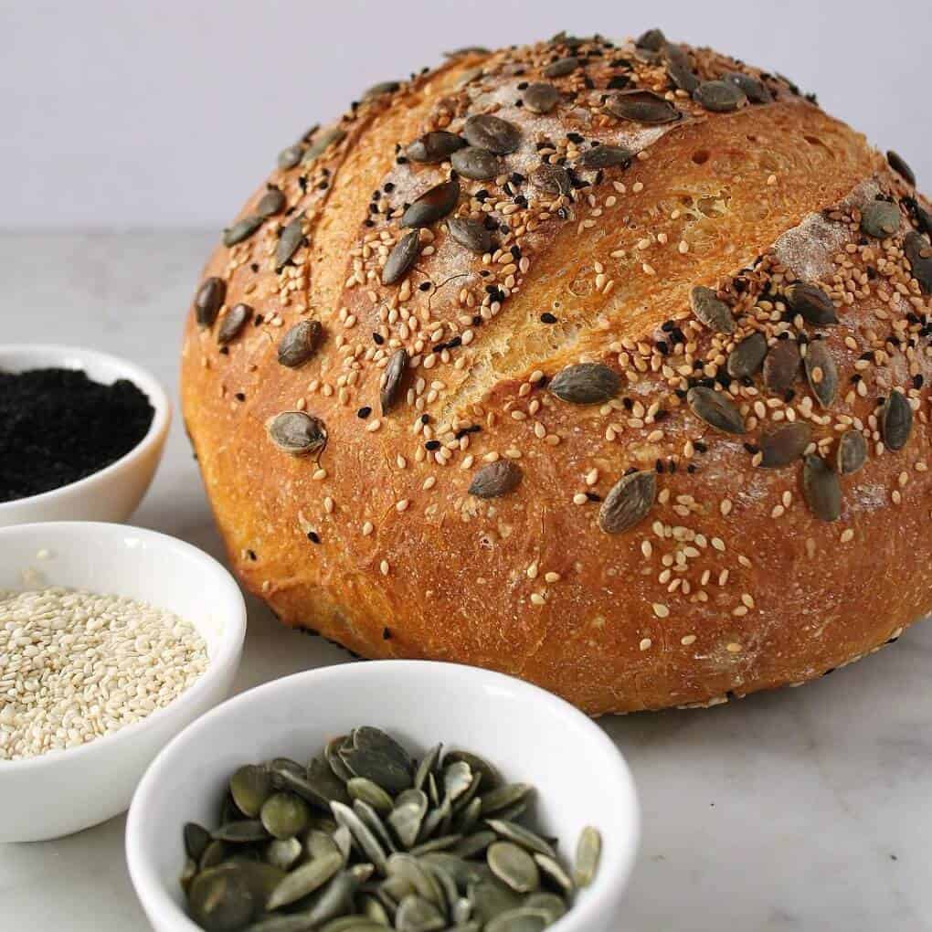 Pumpkin No Knead Bread - Bread boule topped with various seeds, surrounded by small bowls filled with the same seed.