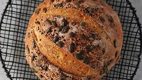 Pumpkin No Knead Bread - Bread Boule topped with assorted seeds on a black cooking rack.