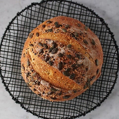 Pumpkin No Knead Bread - Bread Boule topped with assorted seeds on a black cooking rack.