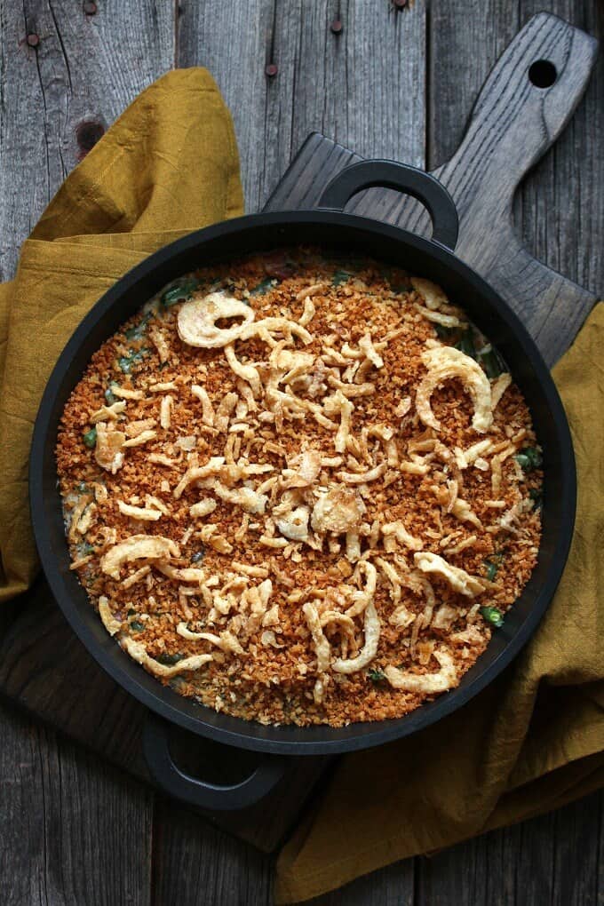 Zesty Green Bean Casserole - An overhead shot of a golden crumb and onion topped casserole in a black baking dish surrounded by yellow napkins on a grey wooden background.