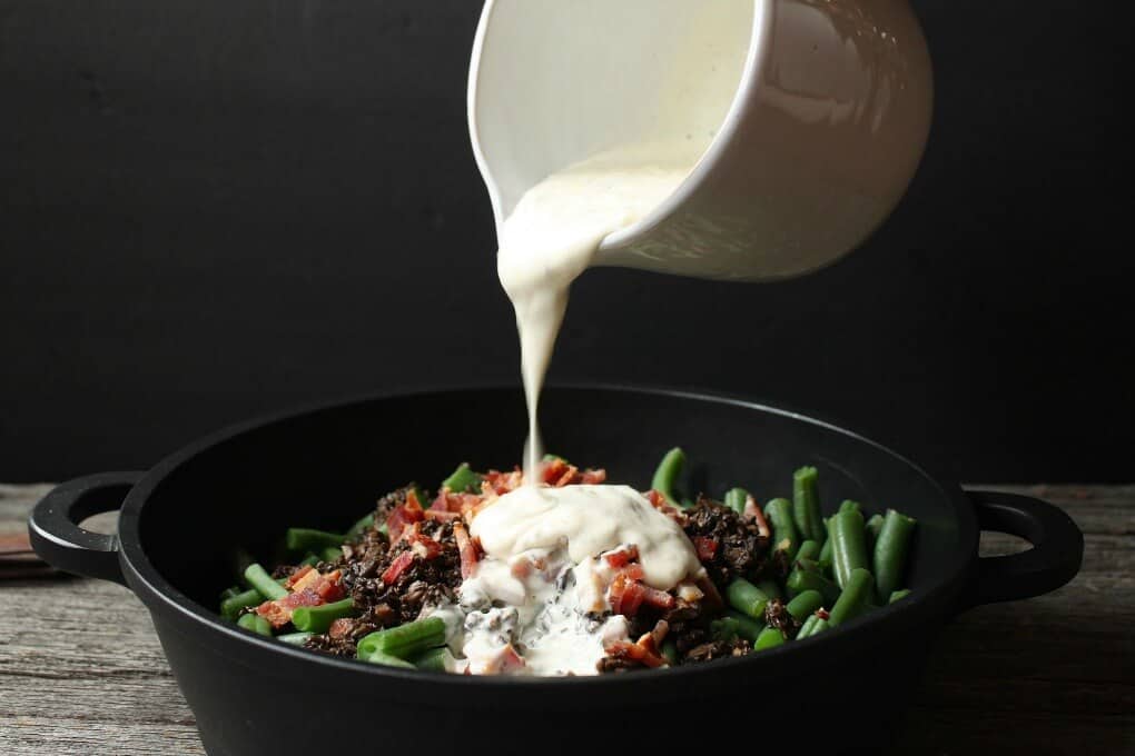 A creamy sauce being poured over the ingredients of a green bean casserole.