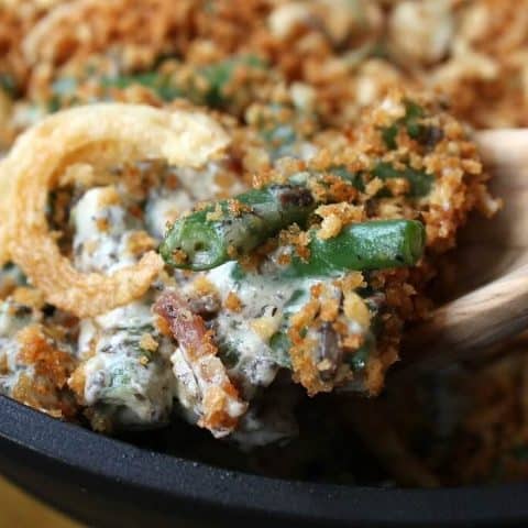 Zesty Green Bean Casserole - Close up of a wooden spoon holding a serving of fried onion, bacon, and green bean casserole.