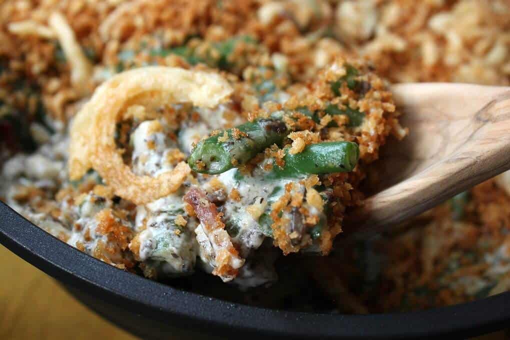 Zesty Green Bean Casserole - Close up of a wooden spoon holding a serving of fried onion, bacon, and green bean casserole.