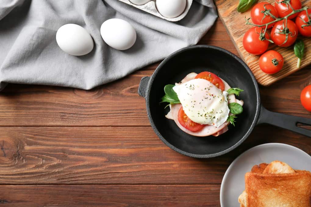 Eggs Benedict in a small cast iron pan with fresh eggs tomatoes and toast surrounding it. 