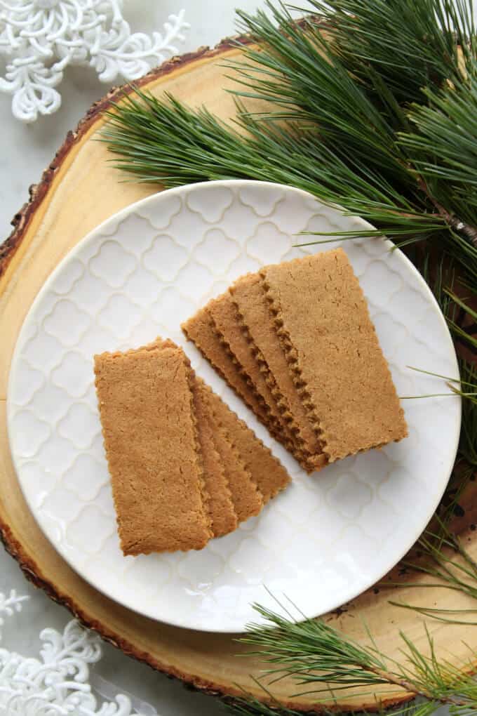 A white textured plate filled with sparkling brown rectangular cookies surrounded by evergeen branches.