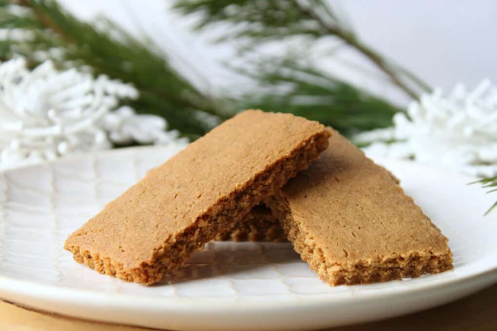 A white textured plate filled with sparkling brown rectangular cookies surrounded by evergeen branches.