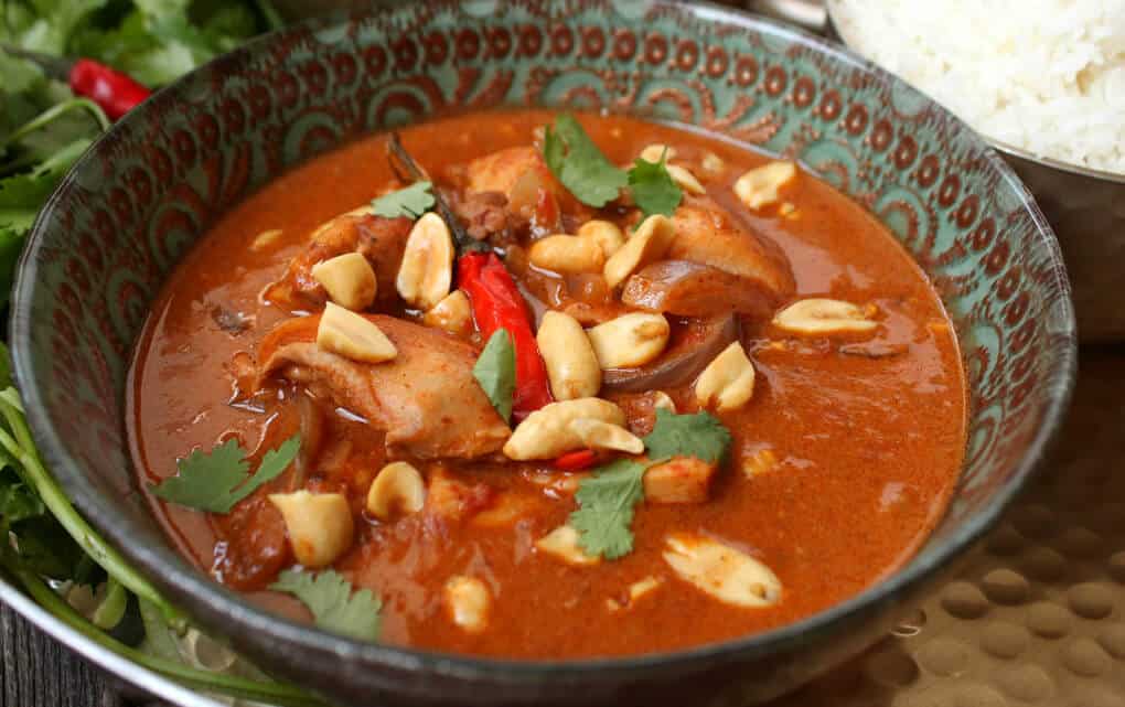 Close up of an ornate bowl filled with rich brown chicken curry topped with peanuts and cilantro.