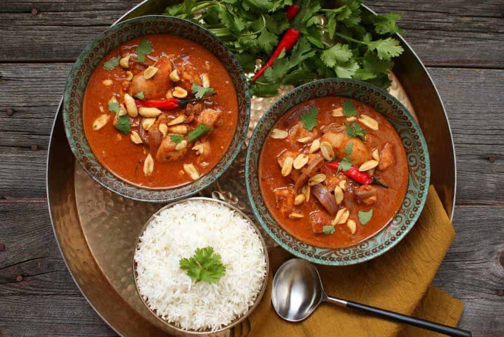 A gold tray holds two bowls of bright brown curry, rice, cilantro and Thai red peppers.