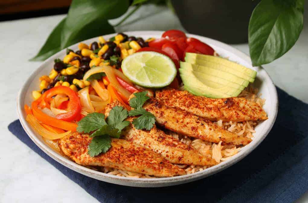 Healthy Chicken Burrito Bowl - A low white bowl filled with healthy vegetables, bean salad, avocado, rice, and chicken tenders. 