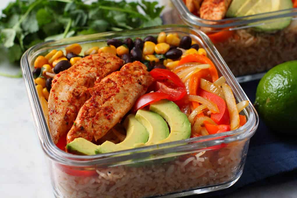 Healthy Chicken Burrito Bowl - Close up of a glass container filled with healthy vegetables, bean salad, avocado, rice, and chicken tenders. 