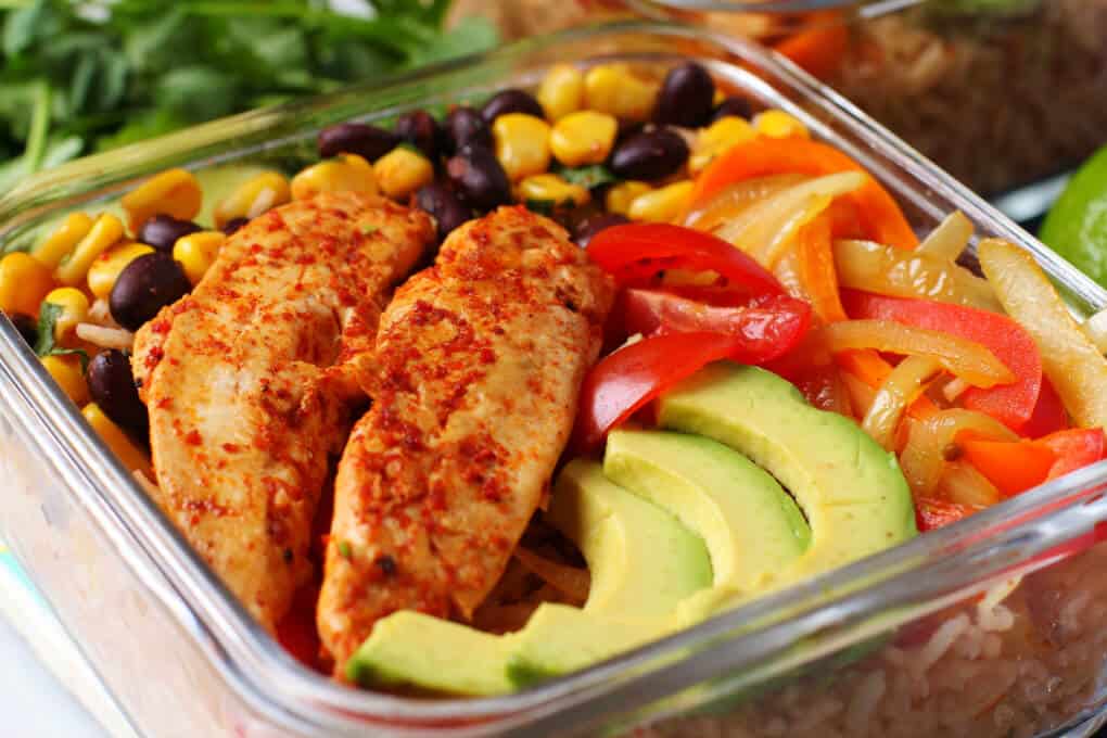 Healthy Chicken Burrito Bowl - Close up of a glass container filled with healthy vegetables, bean salad, avocado, rice, and chicken tenders. 