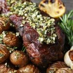 Slow Roasted Boneless Leg of Lamb - close up view of a platter filled with lamb roast, mini hasselback potatoes, lemons, asparagus, and roasted garlic.