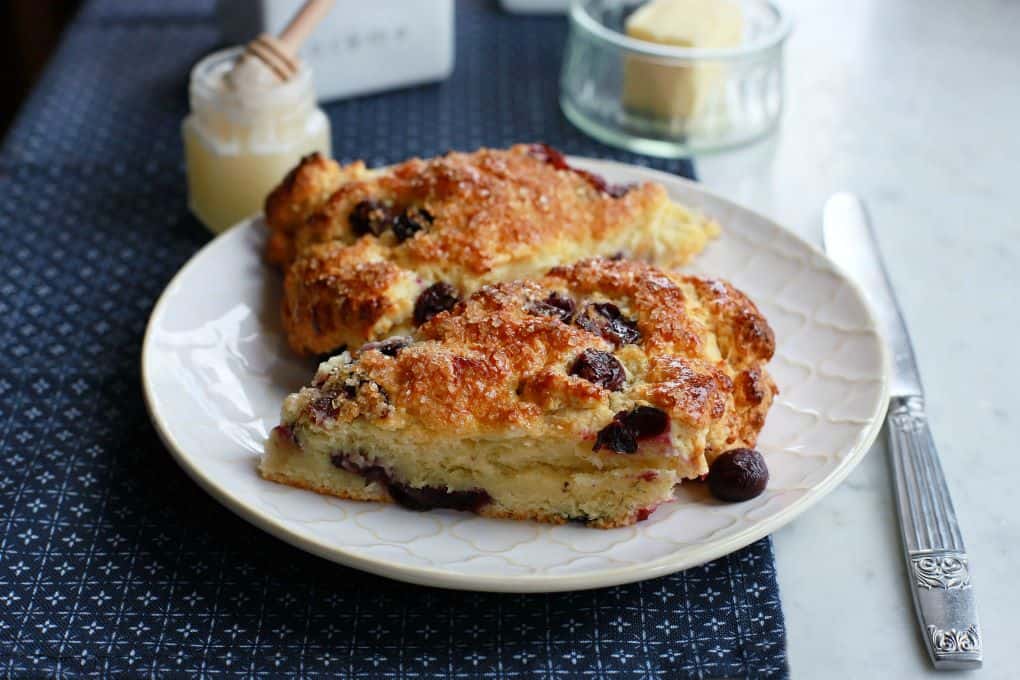 Two golden baked blueberry scones on a white plate surrounded by a butter dish, honey, and cream container. 