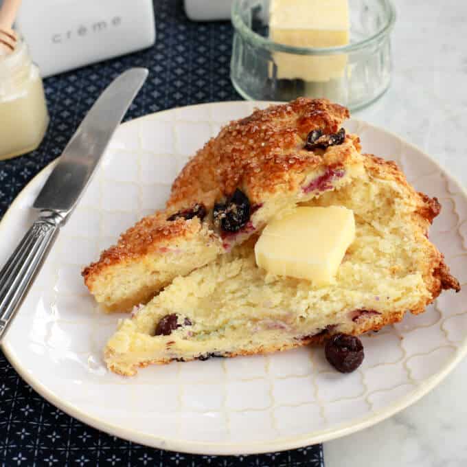 Buttermilk Blueberry Scones - A sliced blueberry scone with a pat of butter on a white plate surrounded by a butter dish, honey, and cream container. 