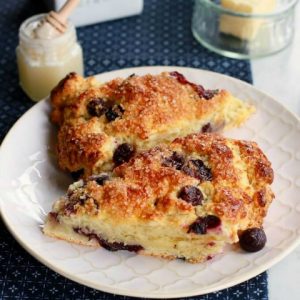 Buttermilk Blueberry Scones - Two golden baked blueberry scones on a white plate surrounded by a butter dish, honey, and cream container.