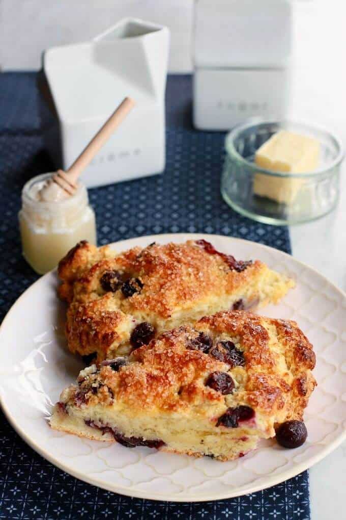 Two golden baked blueberry scones on a white plate surrounded by a butter dish, honey, and cream container. 
