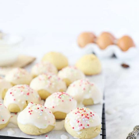 Side view of anise cookies on a baking rack with a white glaze and nonpareils. Eggs in the background