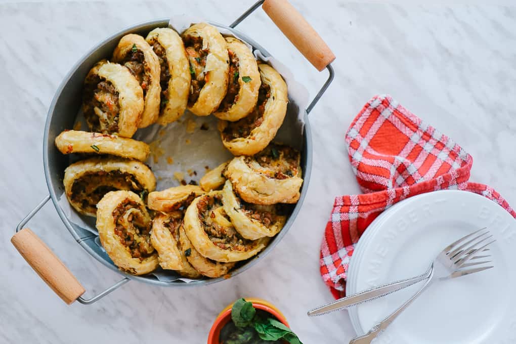 Overhead shot of sausage pinwheels in a metal bowl with handles and white plates with forks