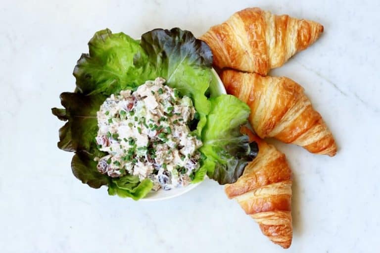 A bowl of chicken salad on a bed of leafy greens next to three croissants, set on a light marble surface. The chicken salad is garnished with chives and contains diced ingredients.