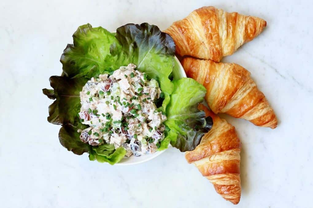 Chicken salad in a lettuce lined bowl with three croissants on a white marble background.